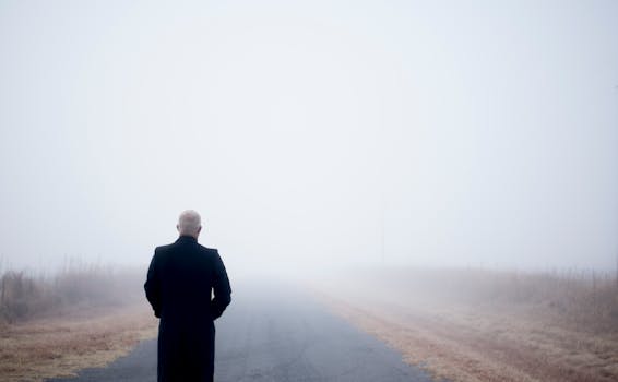 A lone man in a black coat stands on a foggy road, embodying solitude and introspection.
