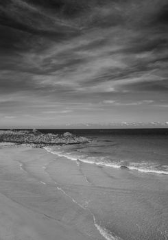 A calming black and white scene of ocean waves gently hitting a quiet sandy beach under a cloudy sky.