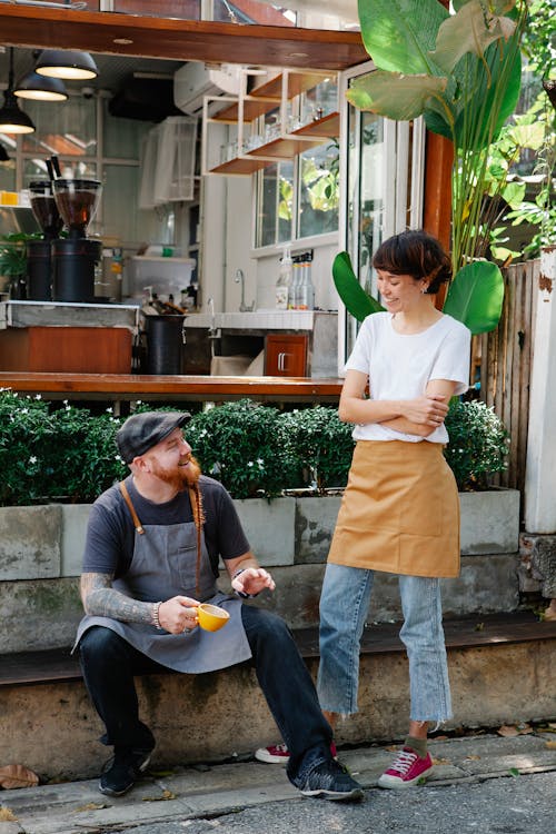 Couple of happy barista colleagues in casual clothes and aprons having conversation in street near cafeteria with cup of drink near plants in daylight and looking at each other