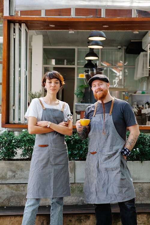 Confident couple of barista coworkers in casual outfit and aprons standing with cup of coffee and portafilter in street near plants and cafe while looking at camera