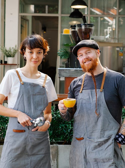 Smiling couple of barista coworkers in aprons and casual outfit spending time in street with portafilter and cup of coffee near cafeteria and plants while looking at camera