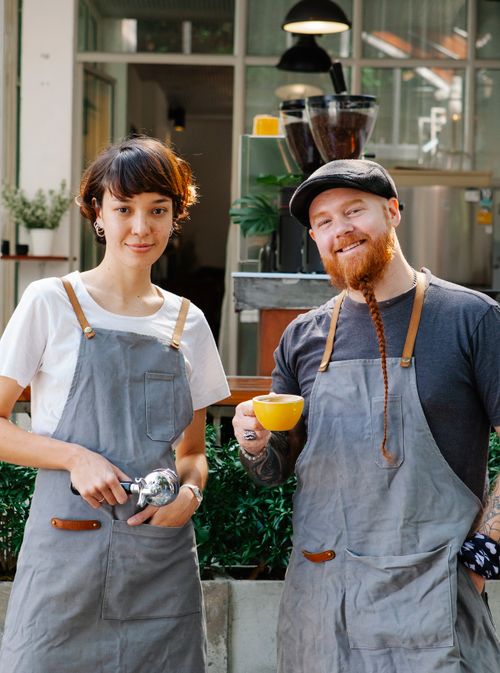 Free Smiling couple of barista coworkers in aprons and casual outfit spending time in street with portafilter and cup of coffee near cafeteria and plants while looking at camera Stock Photo