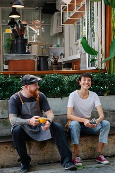 Full body of happy couple of barista colleagues in aprons and casual clothes sitting near cafe in street near plants and having conversation with cup of coffee and portafilter in daylight