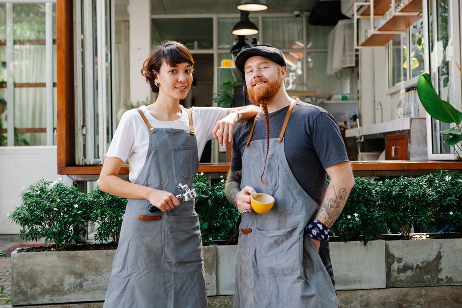 Confident couple of colleagues in aprons and casual clothes standing near cafe in street with portafilter and cup of cof