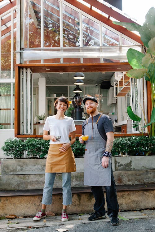 Full body couple of smiling barista colleagues in casual clothes and aprons standing with portafilter and cup of drink in street with plants near cafeteria and looking at camera