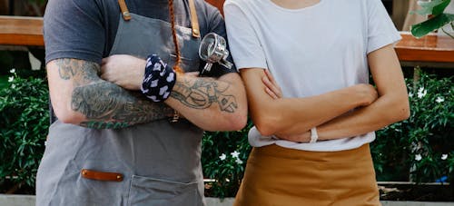 Crop anonymous couple of barista coworkers in aprons and casual clothes spending time in street with portafilter near plants in daytime