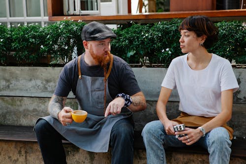 Couple of barista coworkers in aprons and casual outfit sitting near cafe in street and talking with cup of coffee and portafilter near plants in daytime