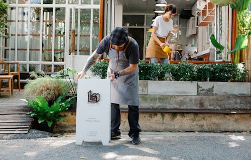 Free Couple of coworkers in aprons and casual clothes watering plants and putting signboard near cafeteria in daytime Stock Photo