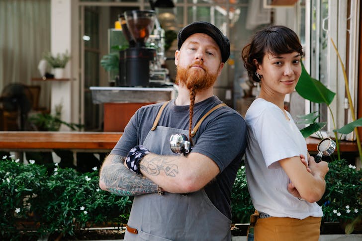 Couple of confident barista coworkers in aprons and casual outfit standing in street near cafeteria with portafilter near plants while looking at camera in daylight