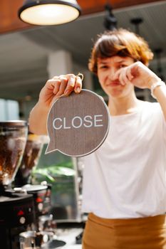 Young female barista in casual outfit and apron standing in light cafeteria and demonstrating small sign with word close while making sad face and looking at camera