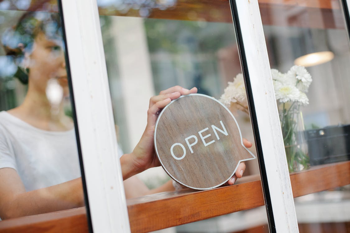 Young woman worker in casual clothes standing in light cafe and putting sign with word open on door