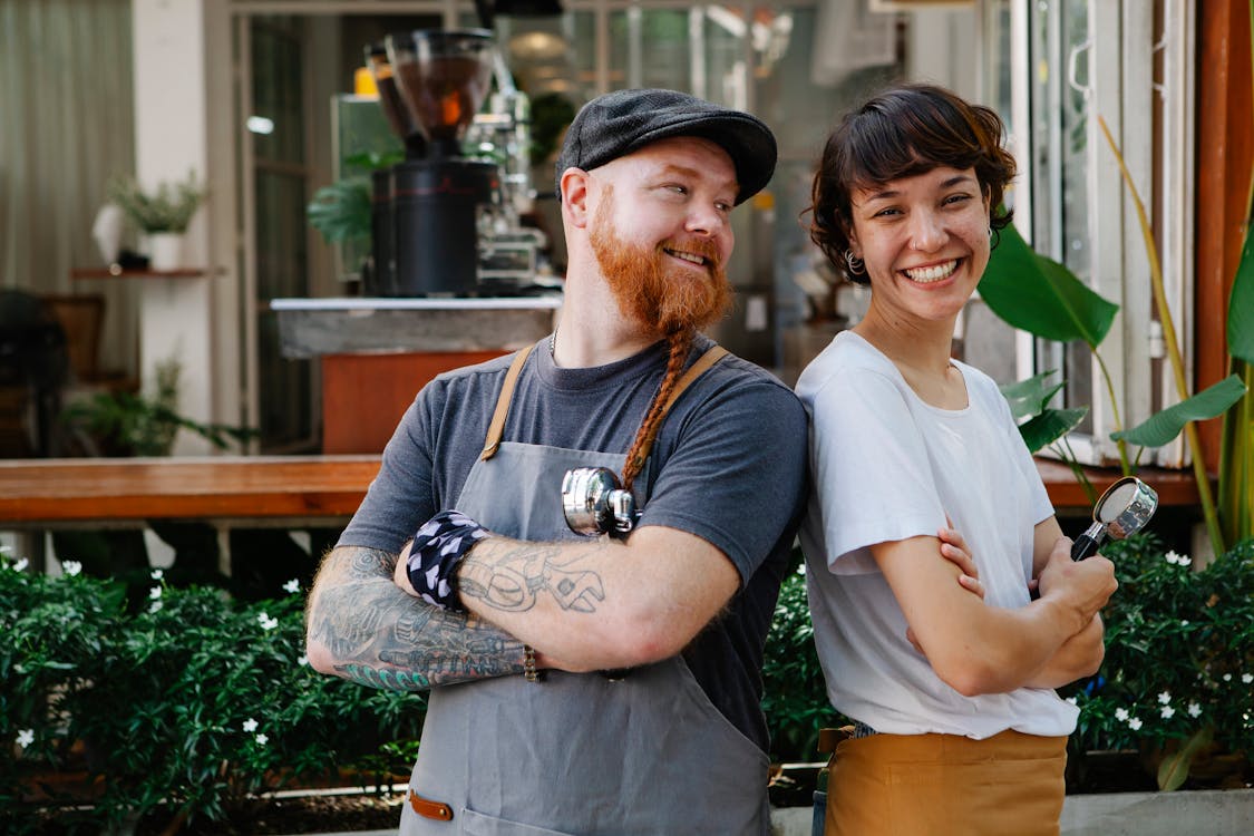 Free Couple of smiling barista coworkers in casual outfit and aprons standing with portafilter near cafeteria in street near plants in daylight Stock Photo