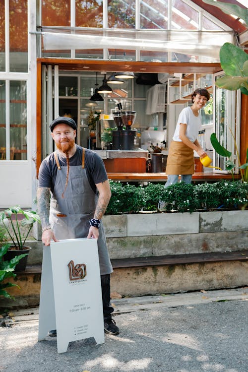 Happy couple of coworkers in aprons and casual clothes standing near signboard and watering plants near cafe in daytime and looking at camera