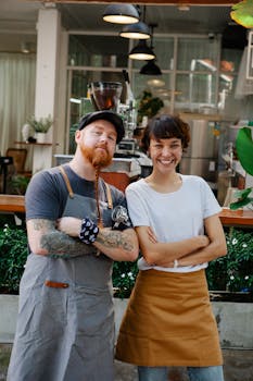 Couple of confident colleagues in casual clothes and aprons spending time in street near cafe with portafilter near plants while looking at camera in daytime