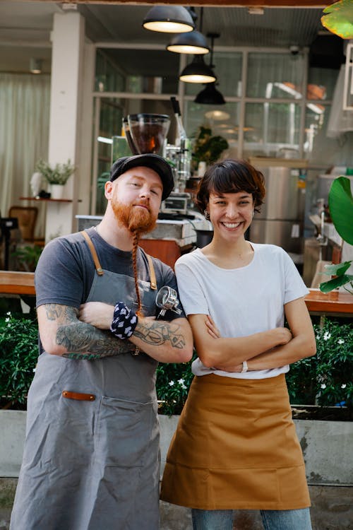 Couple of confident colleagues in casual clothes and aprons spending time in street near cafe with portafilter near plants while looking at camera in daytime