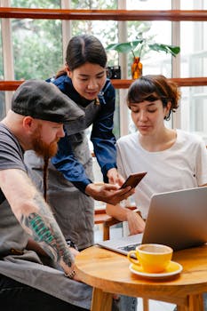 Group of colleagues in aprons and casual clothes using computer and phone in cafe with cup of drink in daylight