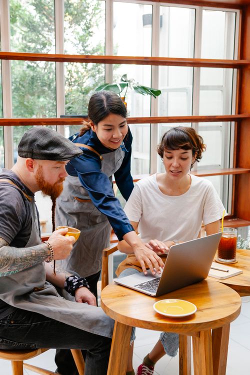 Free Group of coworkers in casual outfit and aprons surfing on laptop in cafeteria at table with cup of drink in daytime Stock Photo