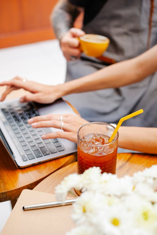 Crop faceless coworkers in aprons using computer at table in light cafeteria with coffee cup and glass of drink near flowers and paper with pen