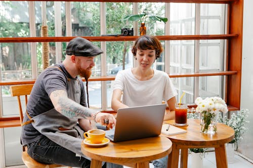 Couple of colleagues in aprons and casual clothes in cafe working on laptop at table with glass of drink and coffee cup in daylight