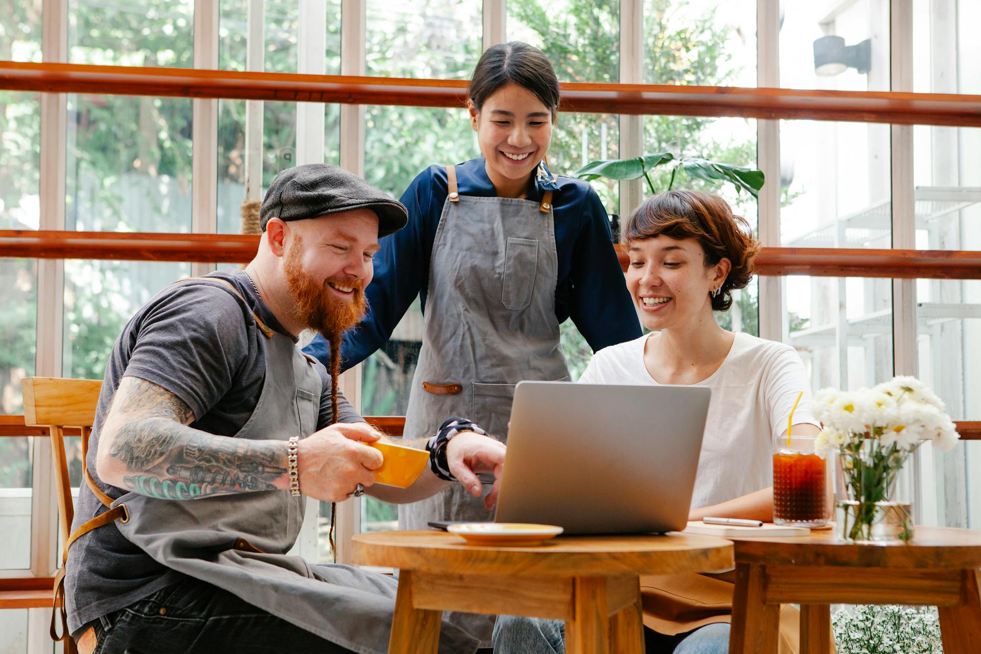 Positive coworkers in aprons and casual outfit using computer in cafeteria at table with coffee cup and glass of drink in day