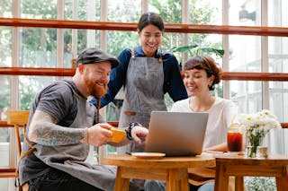 Positive coworkers in aprons and casual outfit using computer in cafeteria at table with coffee cup and glass of drink in day