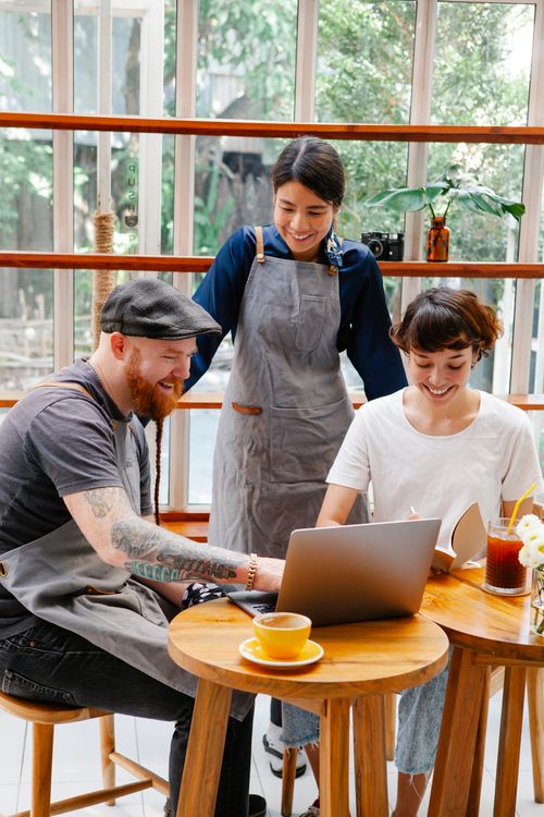 Free Smiling colleagues in casual clothes and aprons in cafe working on netbook at table with cup of drink in daylight Stock Photo