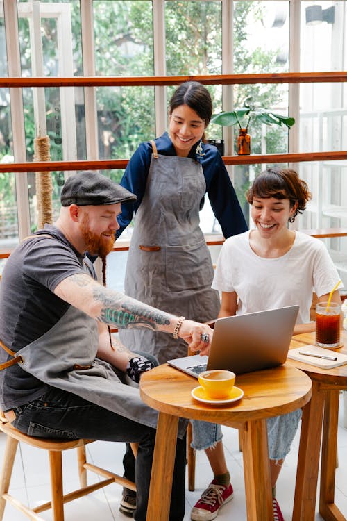 Cheerful multiethnic cafeteria staff and female barista talking at wooden tables with netbook in daylight