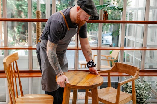 A barista adjusts wooden furniture inside a modern cafe with large windows and plants.
