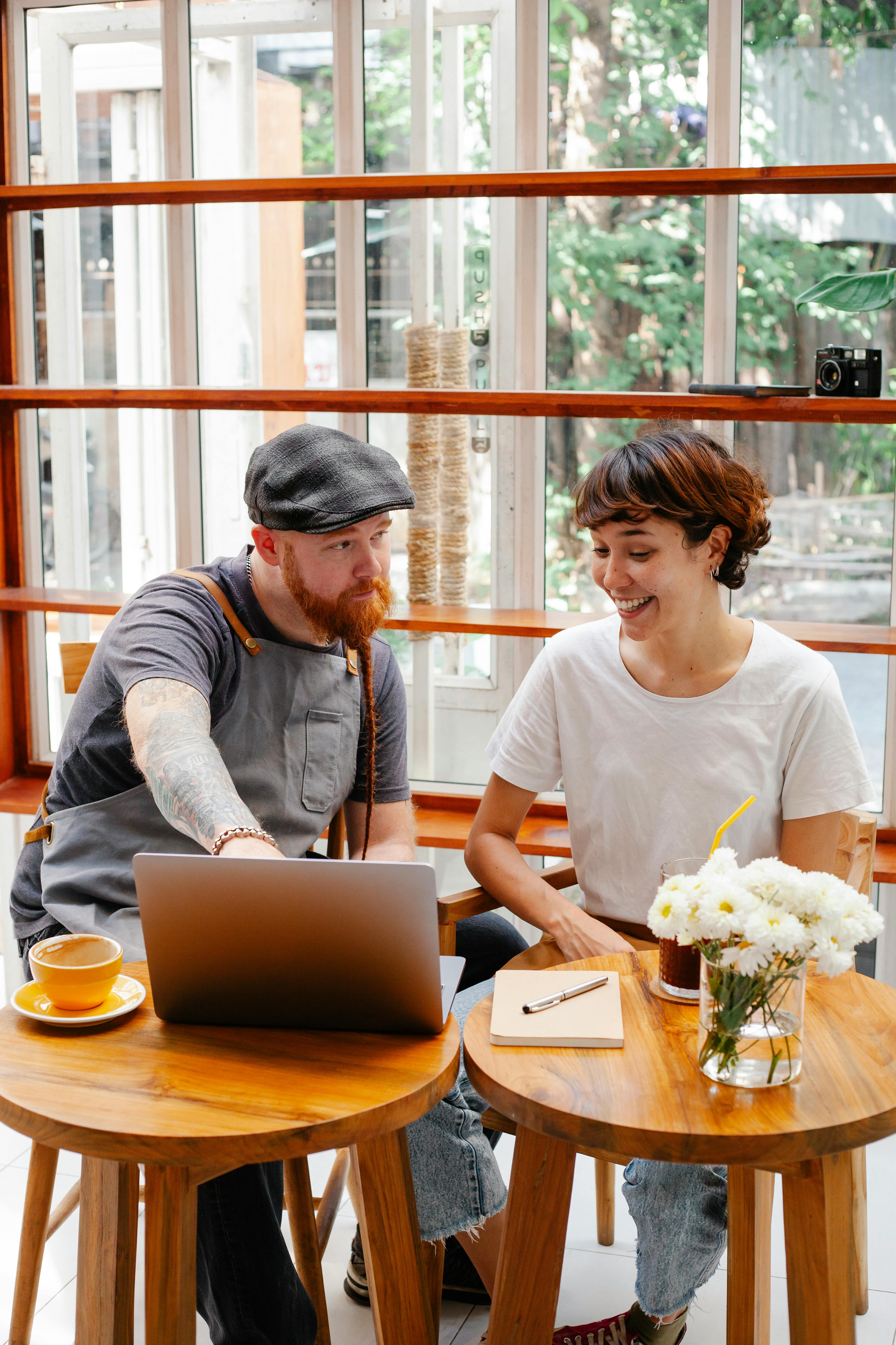 content barista speaking with hipster partner against laptop in cafe