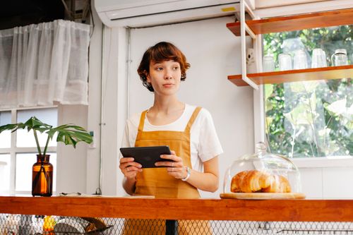 Free Young dreamy female employee in apron with tablet looking away against delicious puffs in bakery Stock Photo