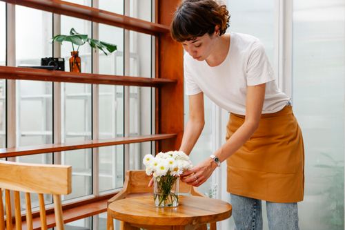 Free Crop young female cafeteria worker putting vase with blossoming flowers on wooden table in daytime Stock Photo