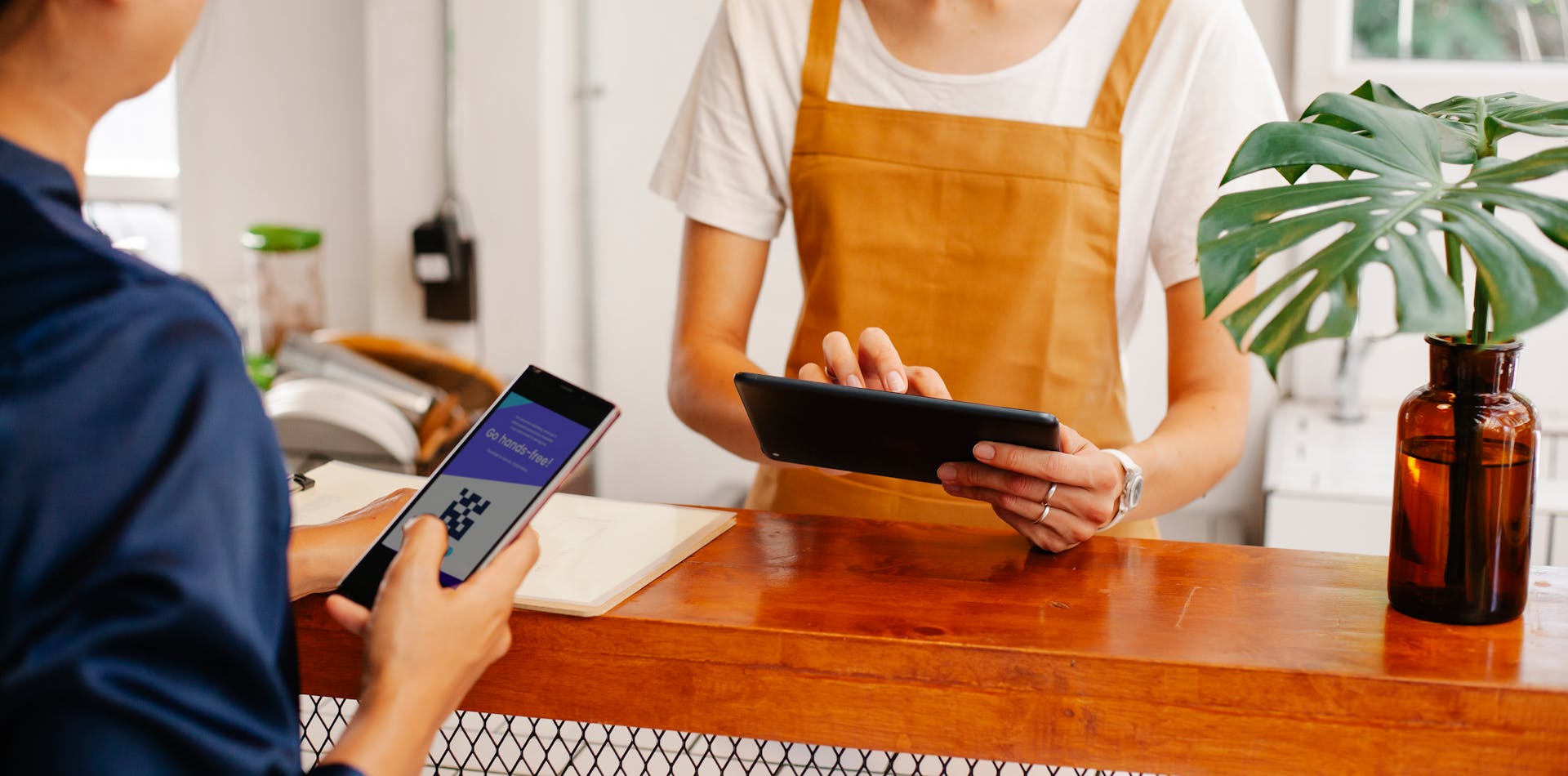 Crop anonymous female employee with application on cellphone screen interacting with partner using tablet at counter in cafeteria