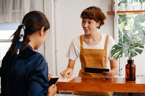 Cheerful young cafeteria worker pointing at paper while speaking with anonymous coworker with smartphone in daytime