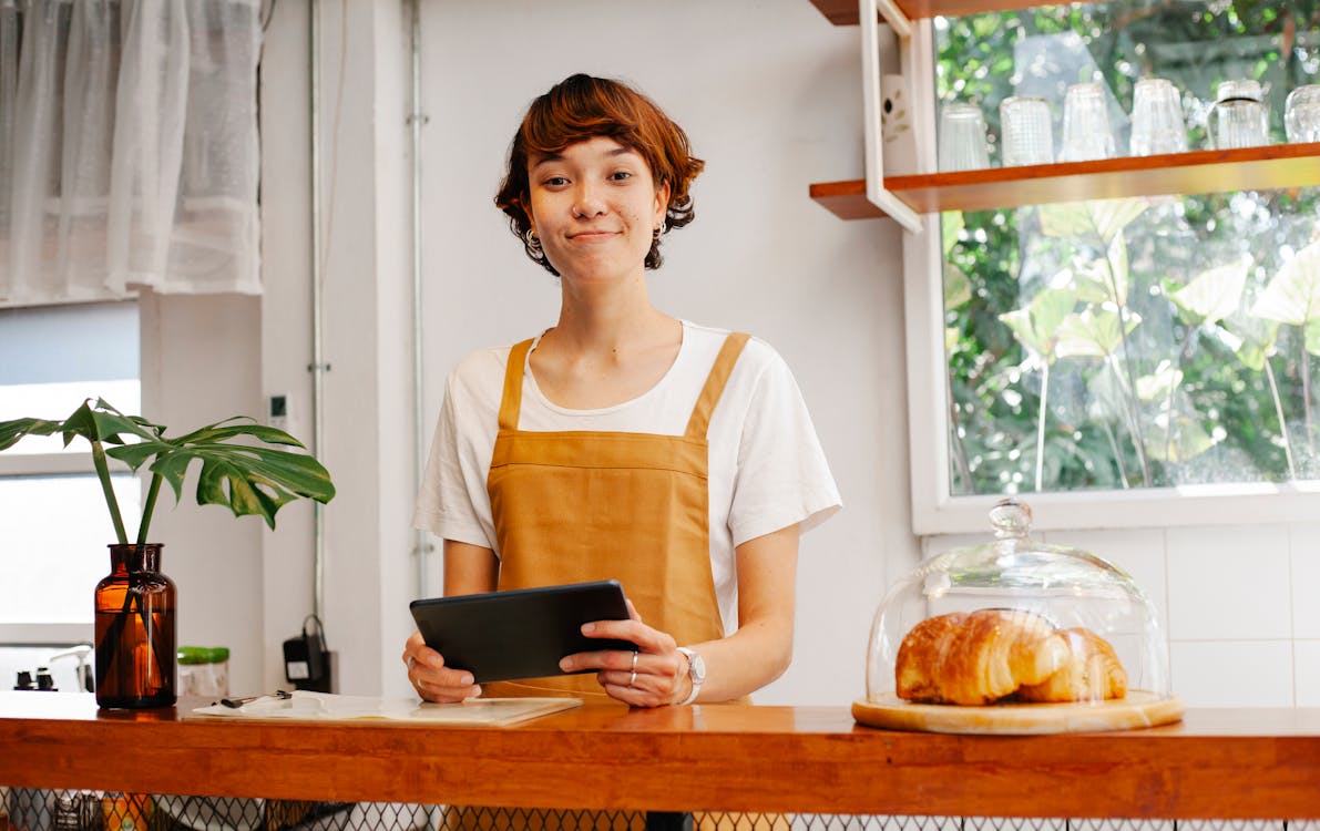 Free Sincere young female cafeteria worker with tablet looking at camera near container with tasty pastries Stock Photo
