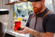 Crop cheerful hipster male barkeeper with transparent glass of beer beverage with garnish on top in bar kitchen