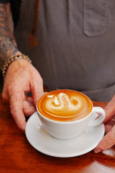 A barista with tattoos carefully holding a cappuccino with latte art on a wooden table.