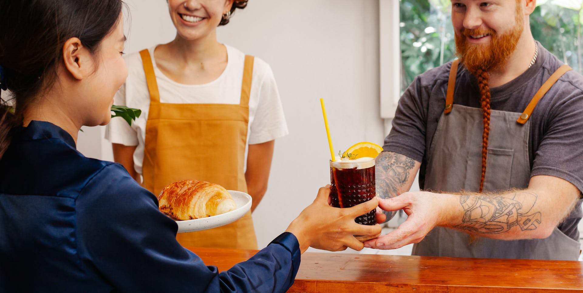 Crop cheerful workers passing tasty pastry and glass of alcoholic beverage to ethnic partner at counter