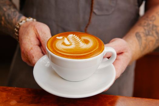 Crop unrecognizable tattooed male employee with cup of tasty coffee with ornament on foam in cafe