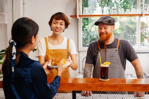 Free Cheerful female worker passing delicious puff to unrecognizable partner above counter near smiling tattooed man Stock Photo