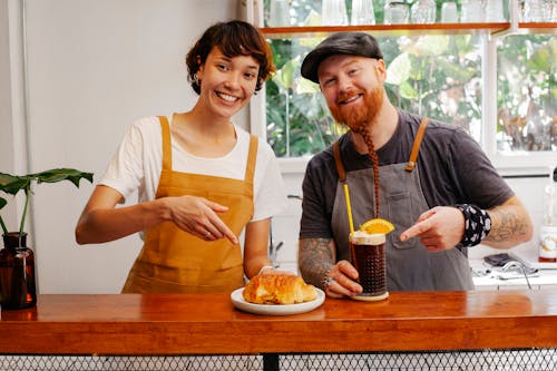 Free Happy coworkers showing appetizing puff on plate and glass of beer cocktail with garnish while looking at camera at counter Stock Photo