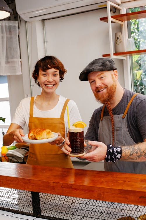 Free Content young female employee with appetizing puff near bearded coworker with beer cocktail looking at camera behind counter Stock Photo