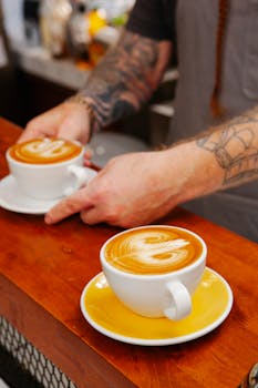 Crop unrecognizable tattooed male cafeteria employee serving cups of tasty latte with ornament on smooth froth