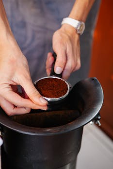 Crop anonymous female cafe employee with aromatic coffee powder in stainless steel portafilter above grinder
