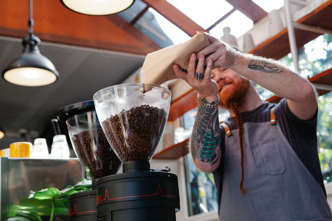 From below of tattooed male employee pouring aromatic coffee beans from paper bag into electric grinder in cafe kitchen
