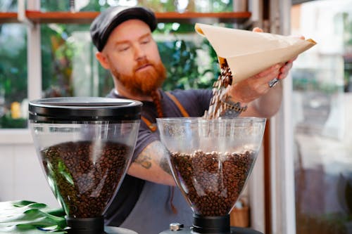 Unshaven employee pouring coffee beans into grinder in cafe