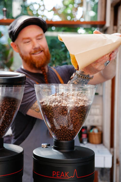 Content adult bearded male cafe worker pouring coffee beans from paper bag into professional grinder in kitchen