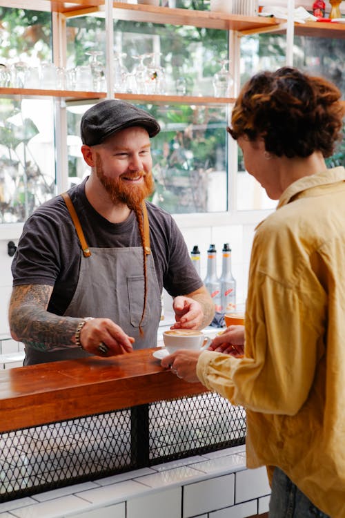 Cheerful hipster barista talking to colleague with coffee at counter