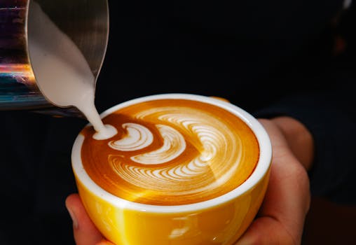 Close-up of milk pouring to create latte art in a yellow cup at a café.