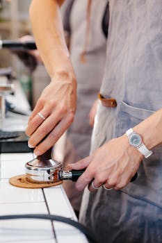 Crop anonymous female employee pressing coffee in metal portafilter with tamper near partner in cafeteria