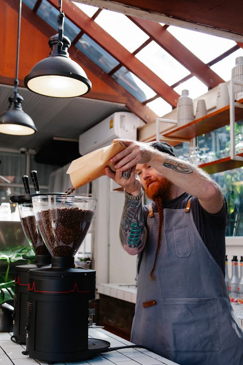 Adult tattooed male employee pouring aromatic coffee beans from paper bag into electric grinder in cafe kitchen